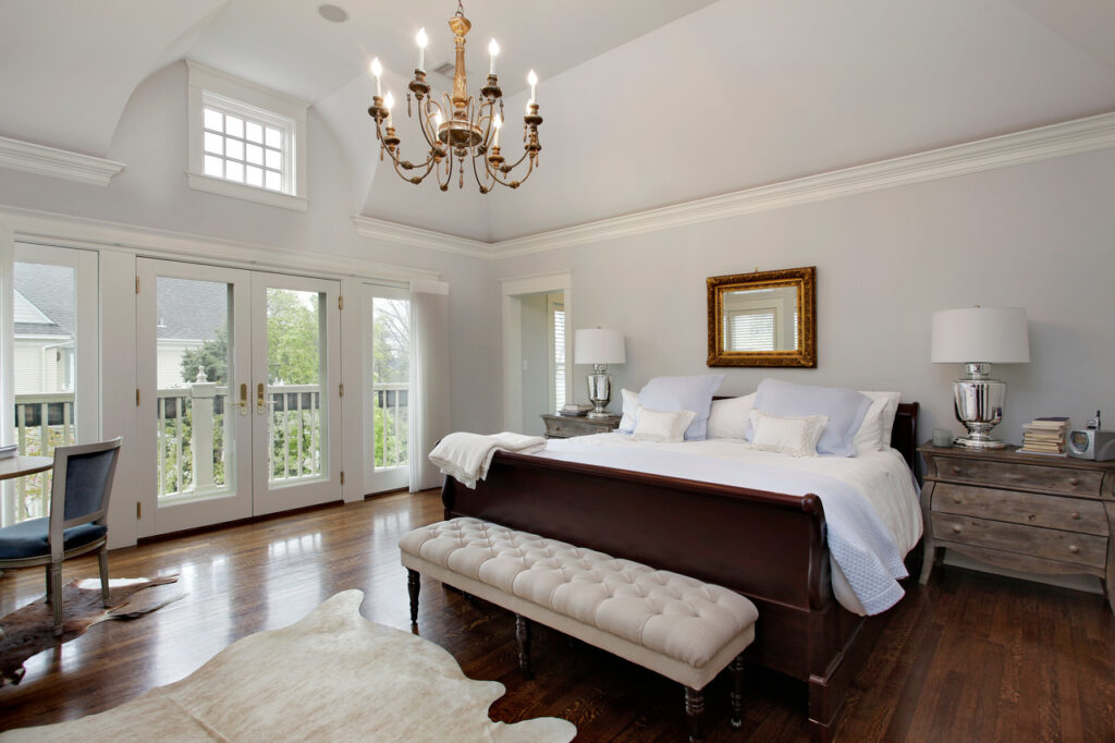 declutter your bedroom Luxurious bedroom with vaulted ceiling, featuring a grand wooden bed with white linens, an upholstered bench at the foot, and elegant chandeliers. The room includes French doors leading to a balcony, gray walls, and a cozy reading nook with a plush chair.