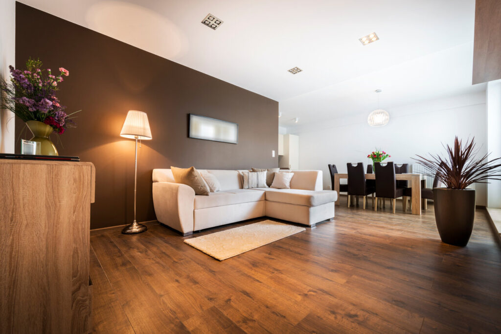 declutter your home Modern living room with light wooden flooring and a white corner sofa set against a brown accent wall. A floor lamp provides warm lighting beside a wooden side table with a vase of flowers.